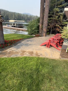 Stone patio and walkway beside a lake before cleaning, the surface pale and dulled, with red Adirondack chairs and a dock behind.