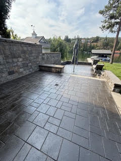 The patio cleared of furniture after sealing, the pavers uniformly dark, with the stone seat wall and the lake behind.